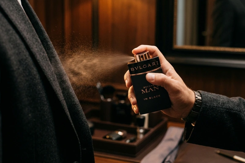 Pessoa aplicando perfume Bvlgari Man in Black em ambiente elegante com fundo de madeira.