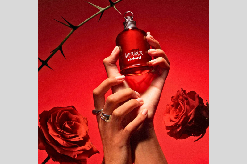 Frasco do perfume Amor Amor da Cacharel cercado por rosas vermelhas, simbolizando paixão e romantismo.