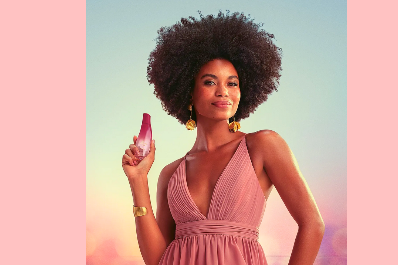 Mulher segurando um frasco de perfume na mão, usando vestido rosa e posando em ambiente com fundo colorido em tons suaves.