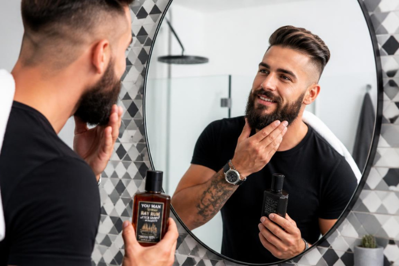Homem barbudo sorrindo e aplicando o melhor pós-barba em frente ao espelho do banheiro.
